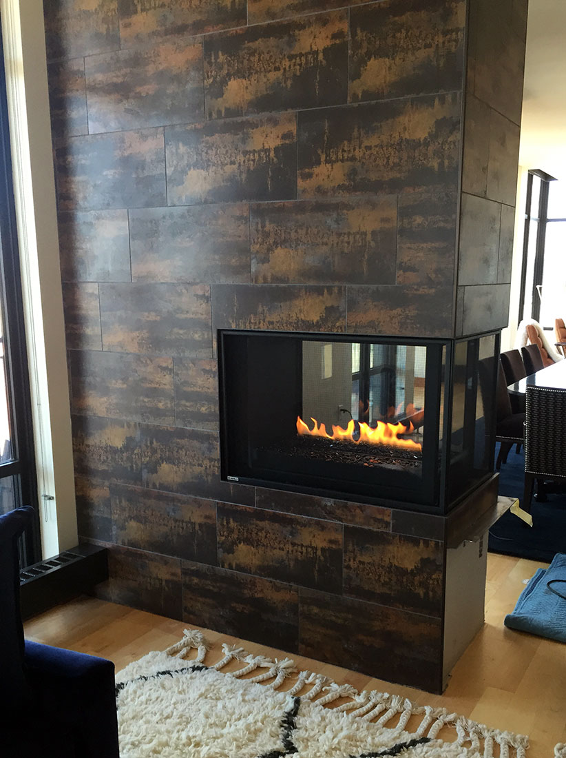 Modern fireplace with a three-sided glass design and rustic tile surround, completed by Carter Custom Construction.